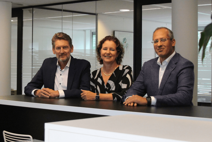 Drie professioneel geklede mensen, twee mannen en een vrouw, zitten glimlachend aan een balie in een modern kantoor met glazen wanden op de achtergrond.