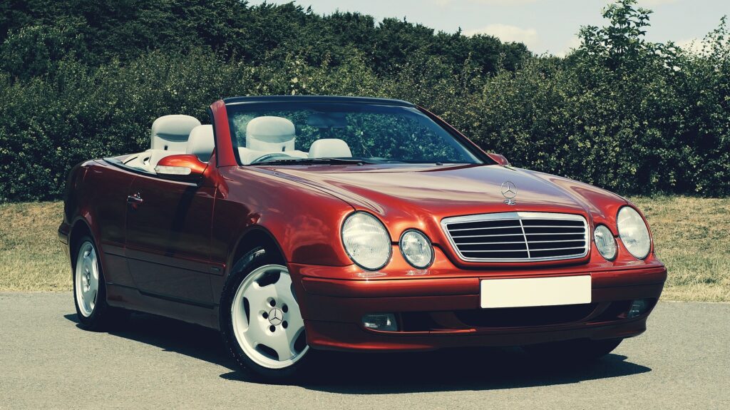 Een glanzend rode Mercedes-Benz cabriolet staat geparkeerd op een weg met groene bomen en struiken op de achtergrond, onder een gedeeltelijk bewolkte hemel.